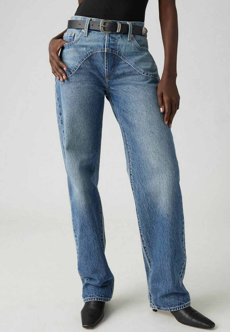 Jean en denim bleu avec une coupe décontractée, taille haute, coutures courbées uniques et ceinture en cuir noir. Texture lisse avec légère décoloration.