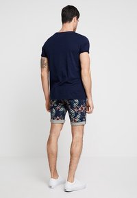 Marinblå t-shirt med rullade ärmar, tillsammans med shorts med blommönster i blått, grönt och rosa, samt uppvikt fåll. Vita sneakers.