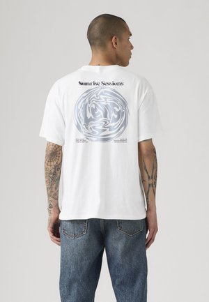 Mand med tatoveringer bærer en hvid T-shirt med abstrakt cirkulært design og teksten "Sunrise Sessions" på ryggen, kombineret med blå jeans.