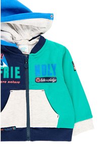 Children's color-block hoodie in navy, teal, and cream with front pockets, zipper closure, hood, and text "kindly take a pledge" on front.