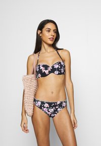 Bikini noir à fleurs avec des fleurs roses et violettes. Le haut est sans bretelles avec un détail froncé ; le bas a une bordure décorative blanche. Portant un sac rose texturé.