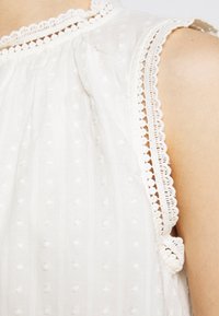 Vit blus med texturerade prickar, spetskant runt halsringningen och ärmhålen, flödande tyg med en mjuk fall.