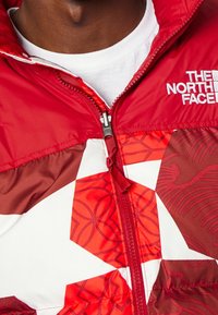 Röd puffjacka med geometriska mönster i nyanser av rött och brunt. Har en hög krage, full dragkedja och vitt logotypdetalj på bröstet.