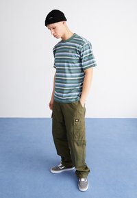 Prugasta kratka majica zeleni, modri in beli; olivno zeleni cargo hlače; črna kapa; sivi superge. Oporne cargo žepe so vidne.