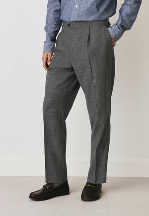 Graue, plissierte Hose aus strukturiertem Stoff, taillierter Schnitt, Bund mit seitlichen Knöpfen verstellbar, kombiniert mit dunklen Lederschuhen.