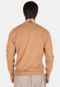Maglione marrone a maglia con maniche lunghe, scollo rotondo e orlo a coste. Texture morbida visibile, indossato con pantaloni grigio chiaro.