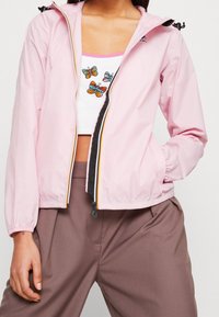 Lyserød letvægtsjakke med elastiske manchetter og frontlynlås. Har en sort lynlås og multikoloriet sommerfuglebroderi på skjorten under.