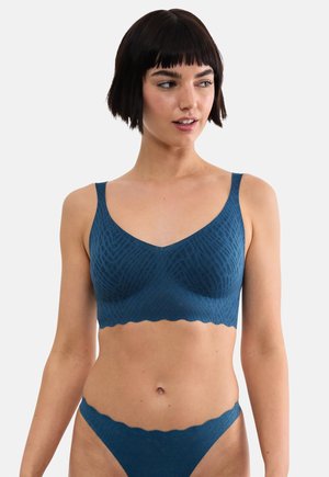 Femme aux cheveux courts et foncés portant un ensemble assorti de soutien-gorge et de culotte bleus sans couture à motif sur un fond uni.