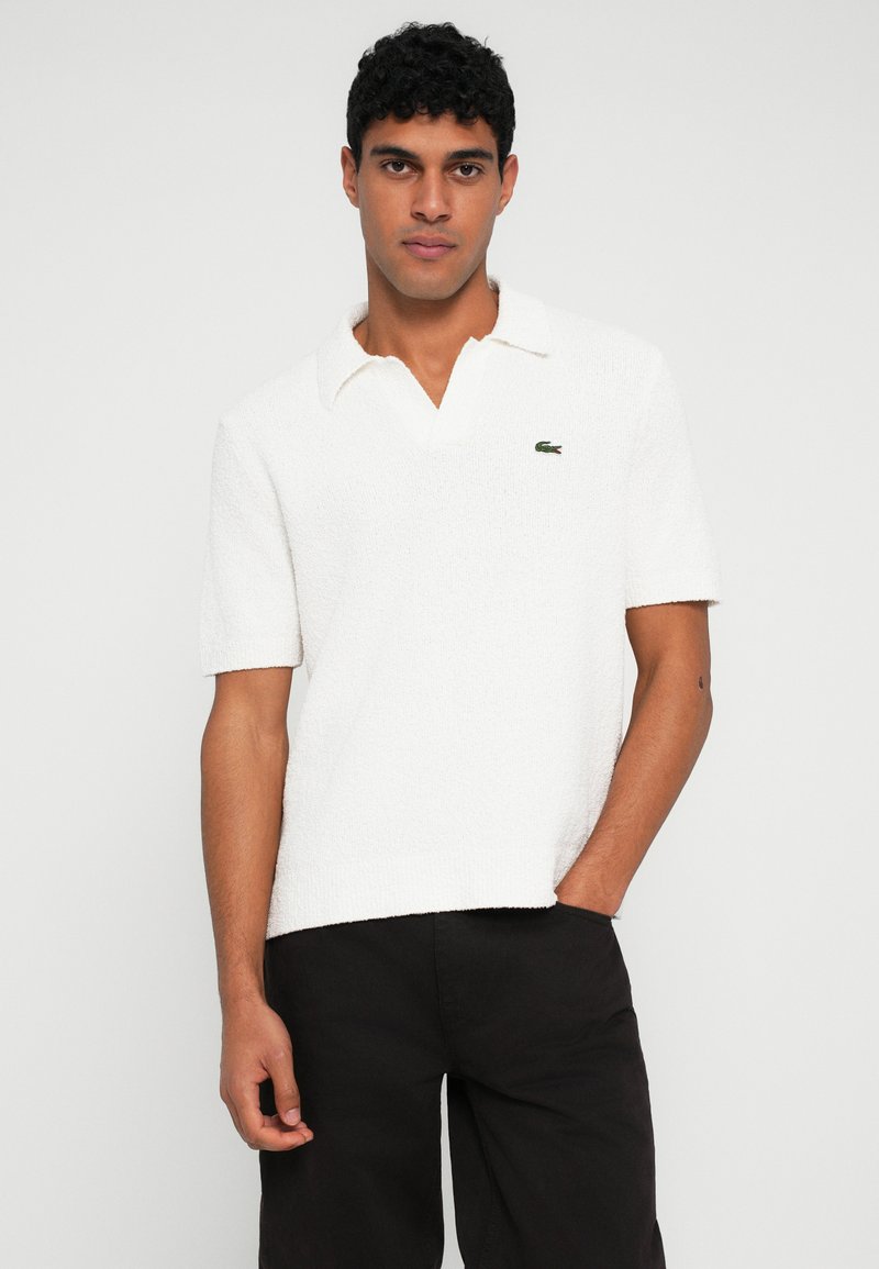 Polo bianco con texture, colletto, maniche corte e logo verde Lacoste sul lato sinistro del petto. Abbinato a pantaloni neri.