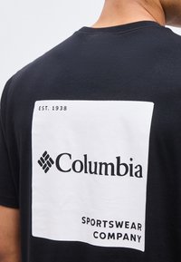 Άτομο που φοράει μαύρο μπλουζάκι με μεγάλο λευκό τετράγωνο στην πλάτη, όπου εμφανίζεται το κείμενο «EST. 1938 Columbia Sportswear Company» και το λογότυπο.