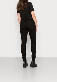 Mujer vestida con jeans ajustados negros, camiseta negra de manga corta y botines negros, de pie sobre un suelo gris claro con un fondo blanco liso.