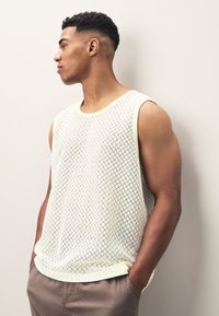 Top in maglia smanicato beige chiaro con motivo a diamante e scollo rotondo. Tessuto liscio con orlo dritto, indossato sopra pantaloni beige.