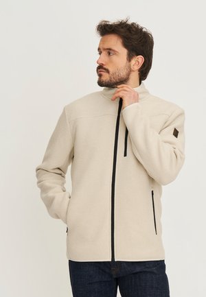 Man in een beige, gestructureerd jack met rits en zwarte ritssluitingen, die met één hand zijn kraag vasthoudt en naar links kijkt.