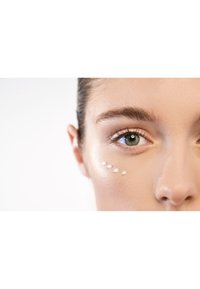 Primer plano de un rostro con una crema beige claro aplicada en pequeños puntos debajo del ojo. La piel tiene una textura suave y luminosa.