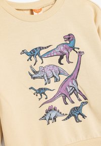 Sudadera beige con un gráfico de varios dinosaurios en azul y morado. El diseño incluye un T-rex, un triceratops y un brontosaurus.