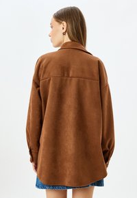 Camicia in finta suede marrone con colletto, maniche lunghe e vestibilità morbida, caratterizzata da un orlo curvo e polsini con bottoni.