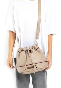 Beige leren emmer tas met paarse accentstrepen, trekkoordsluiting en gouden hardware. verstelbare platte schouderband bevestigd.