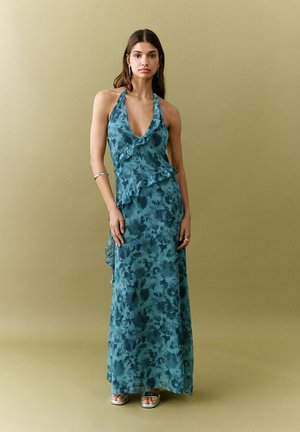 Mujer con un vestido largo sin mangas de cuello halter con estampado floral azul verdoso y volantes, de pie contra un fondo beige liso.
