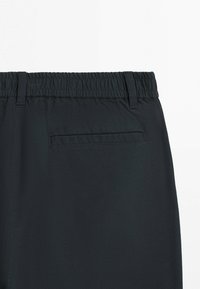 Czarne spodnie z elastycznym pasem, szlufkami na pasek oraz pojedynczą tylnią kieszenią, pokazane na białym tle.