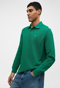 Poloko s dlhým rukávom zelené farby vyrobené z textúrovanej látky, so klasickým golierom a malým logom na ľavej hrudi. V kombinácii s modrými džínsami.