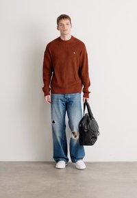 Bruin geribbeld sweater, lichtblauwe versleten jeans, witte sneakers en een zwarte totebag. Eenvoudige, casual outfit met minimale details.