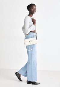 Biała torebka crossbody z klapką i detalem z klamrą. Modelka nosi szary top z długim rękawem, jasnoniebieskie spodnie o szerokich nogawkach oraz czarne buty z ćwiekami.