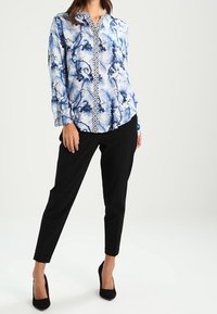 Blouse à imprimé floral bleu avec un fond texturé, devant boutonné et bordure blanche contrastante, associée à un pantalon noir ajusté et des talons.