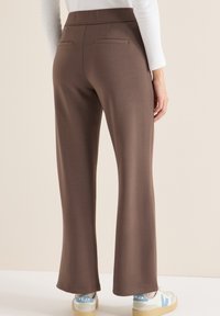 Pantalon droit marron porté par une personne debout, associé à des baskets blanches et un haut à manches longues blanc, sur un fond neutre.