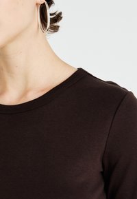 Közelkép egy személy nyakáról és válláról, aki sötétbarna felsőt visel és nagy ezüst karika fülbevalót, rövid, göndör hajjal.