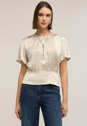 Donna che indossa una blusa beige in raso con maniche corte e vita arricciata, jeans blu a vita alta, accessoriata con una collana d'argento.