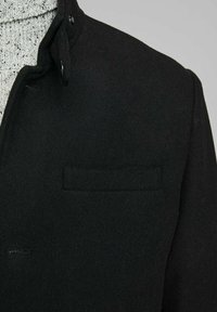 Melnas vilnas maisījuma mētelis ar augstu apkakli, vienu krūšu kabatu un gludu tekstūru. Ar pogu detaļu pie apkakles.