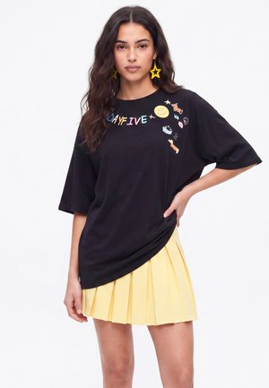 Kvinde med langt mørkt hår iført sort oversized t-shirt med farverige designs og gul plisseret nederdel, poserer med den ene hånd på hoften.