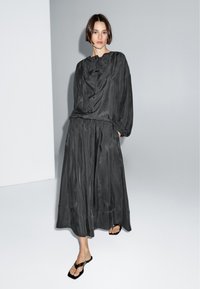 Vrouw die een loszittende zwarte top met lange mouwen en bijpassende broek met wijde pijpen draagt, poseert blootsvoets tegen een effen grijze en witte achtergrond.