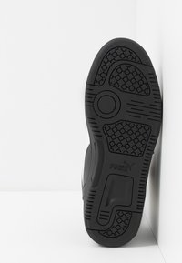 Black rubber sole with circular and hexagonal tread patterns on a white Puma sneaker, positioned upright against a white corner wall.