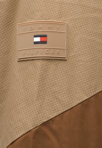 Μπεζ ύφασμα με σχέδιο πλέγματος και καφέ λεπτομέρεια. Μία ταμπέλα από καφέ καουτσούκ με το λογότυπο "TOMMY HILFIGER" και σχέδιο με μπλοκ χρωμάτων.