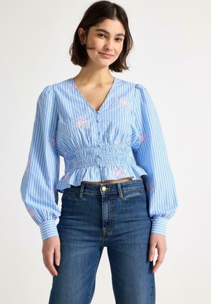 Giovane donna che indossa una camicetta azzurro chiaro con ricami floreali rosa e righe verticali, abbinata a jeans blu scuro a vita alta.