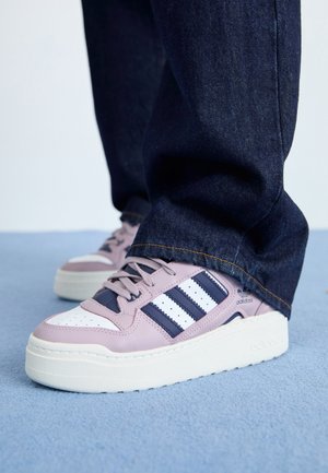 Osoba nosząca różowo-biało-granatowe buty Adidas z trzema paskami, w połączeniu z ciemnoniebieskimi dżinsami, stojąca na niebieskim dywanie.