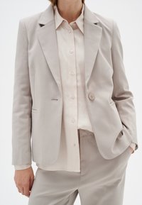 Šviesiai beige pritaikytas švarkas su vienu mygtuku, notch tipo apykaklėmis ir sklandžiu paviršiumi. Nešiojamas virš šviesiai grietinėlės spalvos marškinėlių su mygtukais.