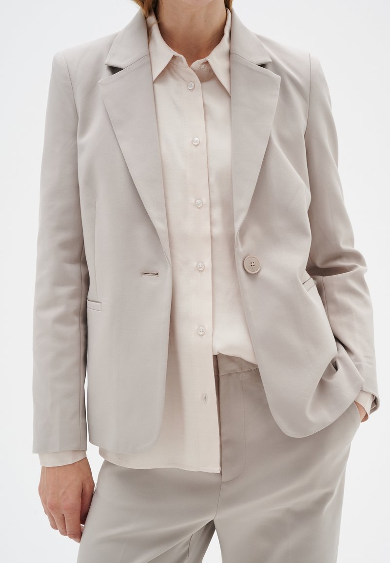 Helle beigefarbene, taillierte Blazerjacke mit einem einzelnen Knopf, Spitzkragen und einer glatten Textur. Getragen über einem hellen cremefarbenen Hemd mit Knöpfen.