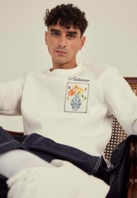 Sweatshirt blanc avec des broderies florales sur la zone de la poche avant, avec des accents bleus. Texture douce, col rond et coupe décontractée.