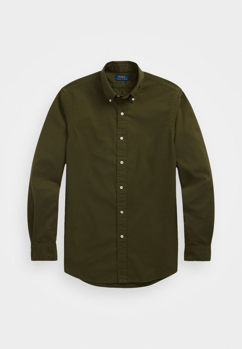 Tamsiai alyvuogių žalios spalvos mygtukų marškiniai su klasikiniu apykakle, ilgomis rankovėmis ir baltais mygtukais. Krūtinėje yra mažas siuvinėtas logotipas.