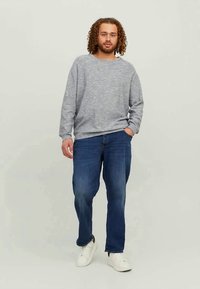 Jovem com cabelo encaracolado, vestindo um moletom cinza, jeans azuis e ténis brancos, de pé com uma mão no bolso, contra um fundo simples.