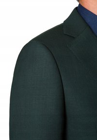 Ciemnozielony żakiet z teksturowanej tkaniny, z wyraźnymi ramionami i ostrym klapem. Niebieski kołnierzyk koszuli wystaje spod kurtki.