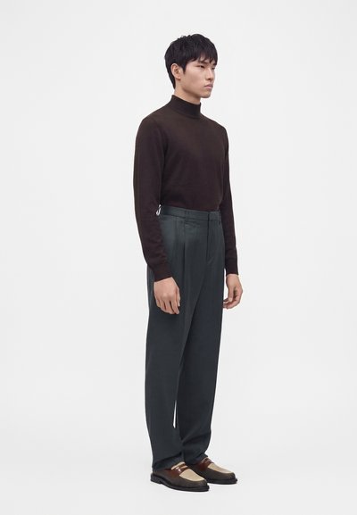 Homme portant un col roulé marron foncé, un pantalon taille haute gris foncé et des mocassins en cuir tressé marron, debout devant un fond blanc.