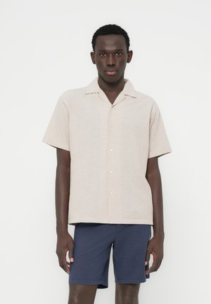 Beżowa koszula na krótki rękaw z kołnierzykiem i guzikami, zestawiona z granatowymi szortami. Koszula wykonana jest z teksturowanej tkaniny i ma luźny krój.