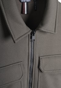 Chaqueta gris con tela texturizada, cierre de cremallera en el frente, cuello puntiagudo y dos bolsillos con solapa. La etiqueta incluye "Antony Morato" y detalles del tamaño.