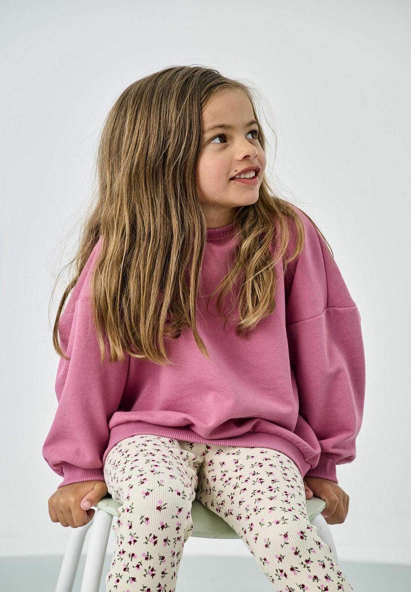 Un sweat-shirt rose à col rond et manches bouffantes, associé à un pantalon crémeux à côtes avec un petit motif floral. Fille assise sur une chaise.