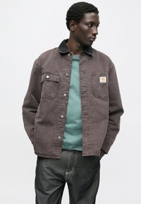 Hnědá džínová bunda s černým manžestrovým límcem, předními kapsami a stříbrnými knoflíky, navrstvená přes tyrkysovou košili a tmavé džíny.