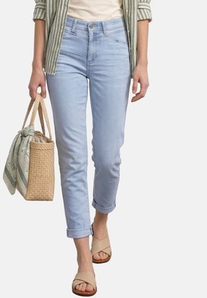 Mujer con vaqueros azul claro remangados, sandalias beige con tiras cruzadas, camisa de rayas y llevando un bolso tejido con pañuelo estampado.