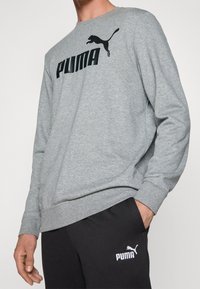 Personne portant un sweat-shirt gris Puma et un pantalon de survêtement noir Puma, debout avec une main dans la poche sur un fond uni.